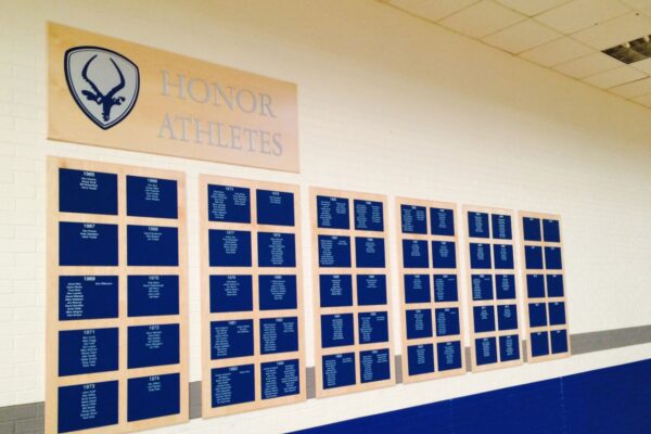 Wall display titled "Honor Athletes" showcasing numerous nameplates organized in a grid, each representing individual athletes. The background is a gymnasium with blue trim on the lower part of the wall. The upper part of the display features an emblem.