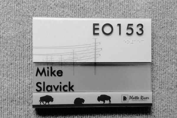 Office nameplate on a gray fabric background, featuring the text "EO153" and "Mike Slavick." The design includes illustrations of power lines and three buffalo silhouettes at the bottom. The Platte River logo is also visible.