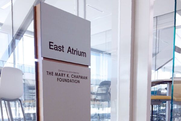 A sign in a modern indoor space reads "East Atrium" and "The Mary E. Chapman Foundation." The background shows a glass doorway with reflections and chairs along the left side. The polished floor reflects parts of the surroundings.