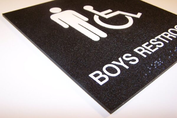 A black, textured sign with white icons indicating a boys' restroom. The sign includes a male figure and a wheelchair symbol, with the text "BOYS RESTROOM" written below. The sign is slightly angled, showing the texture clearly.