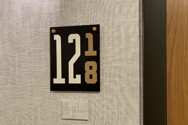 A wall-mounted room number sign reads "1218," with a unique design where the number "1" is smaller and raised above the "2" in the sequence. Below the sign, there's a smaller rectangular plaque displaying "1218" in Braille. The wall has a textured pattern.
