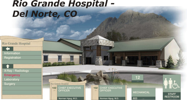 A modern hospital building with a green roof and stone accents, located in front of a mountain backdrop under a cloudy sky. A sign displays services including emergency, surgery, and radiology. Separate directional signs highlight office locations and restrooms.