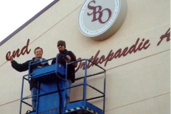 Two workers in winter clothing are standing on an elevated platform. They are smiling and waving while installing or servicing signage on a building that reads "Orthopaedic A..." The building also displays a circular logo with the letters SBO.