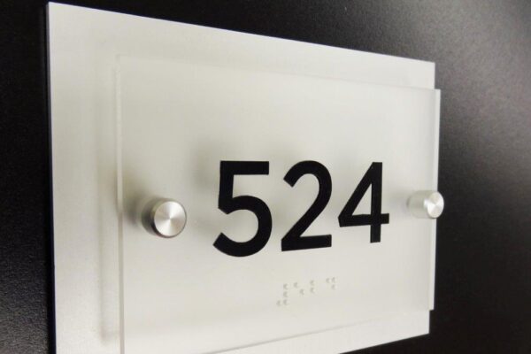 A room number plaque against a black background displays the number "524" in large black font. The plaque is made of frosted glass with two metal bolts securing it to the wall, and includes Braille text below the number.