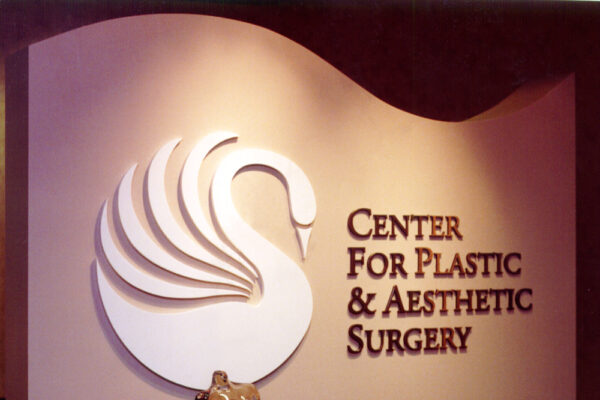 Sign for the "Center for Plastic & Aesthetic Surgery" displayed on a wall. The sign features a stylized white swan logo on the left and the center's name in dark letters on the right. An abstract metallic sculpture is placed on a shelf below the sign.