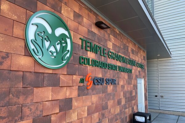 Exterior wall of the Temple Grandin Equine Center at Colorado State University. The wall features the CSU Rams logo and the facility's name in green text, along with the CSU Spur logo. A bench and trash can are visible near the entrance.