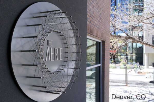 A circular metal sign displaying "The Aerie" is mounted on the side of a building. The sign features a radial design with a geometric pattern. In the background, there are urban buildings and a partial view of tree branches. Text at the bottom reads, "Denver, CO.