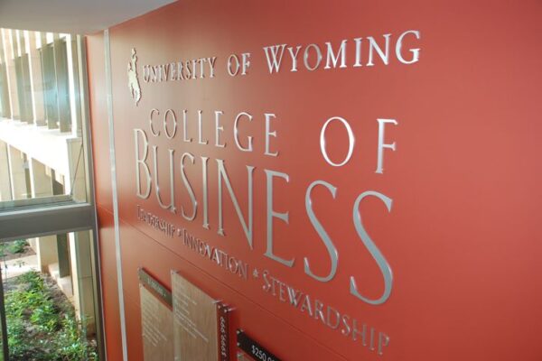 Sign on a red-orange wall reading "University of Wyoming College of Business" with smaller text below it. The sign is located inside a building with glass windows that let in natural light. An informational display case is visible beneath the main text of the sign.