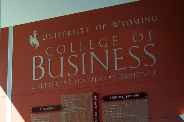 A person stands in front of a large, red wall display at the University of Wyoming College of Business. The display features text about "Leadership," "Innovation," and "Stewardship" along with several panels of detailed information. Light pours in from the left.