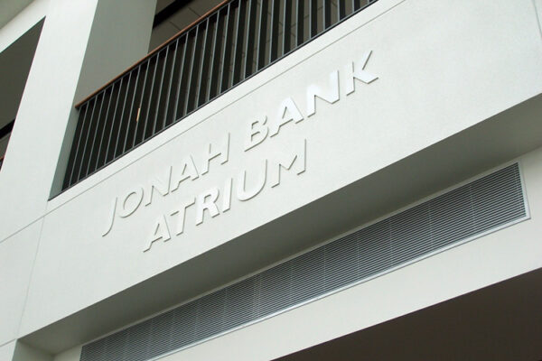 A white concrete wall with the text "JONAH BANK ATRIUM" embossed on it in uppercase letters. The wall has a black horizontal railing near the top.