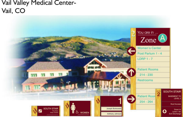 Image showing direction signs and the exterior of Vail Valley Medical Center in Vail, CO. Signs provide navigation to various areas such as Women's Center, Patient Rooms, Restrooms, and exit routes. A picturesque mountain landscape provides a scenic background.