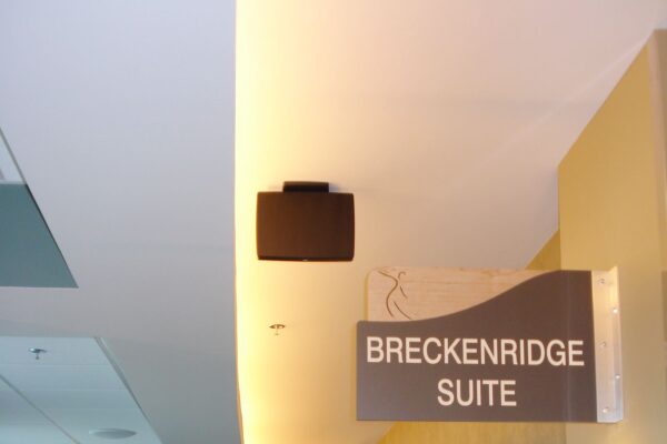 A hallway with modern decor and several seating areas lit by floor and wall lamps. A sign hanging from the ceiling reads "BRECKENRIDGE SUITE." Large windows along the left wall let in natural light. The atmosphere appears calm and inviting.