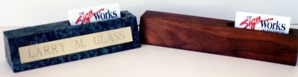 Two nameplate holders are displayed on a surface. The holder on the left is marble with a golden nameplate reading "LARRY M. GLASS." The one on the right is wooden with a slot holding a business card. Both nameplate holders also hold business cards from "The Sign Works.