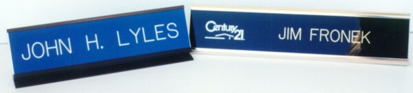 Two nameplates are shown. The one on the left is labeled "JOHN H. LYLES" in white text on a blue background. The nameplate on the right displays "JIM FRONEK" in white text on a blue background, with a stylized logo above the name.