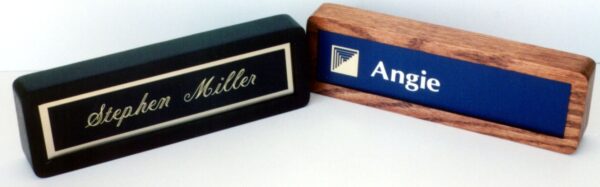 Two desk nameplates are displayed side by side. The left nameplate is black with a gold border and frame, featuring the name "Stephen Miller" in elegant, cursive writing. The right nameplate has a wooden frame and blue background with the name "Angie" and a logo.
