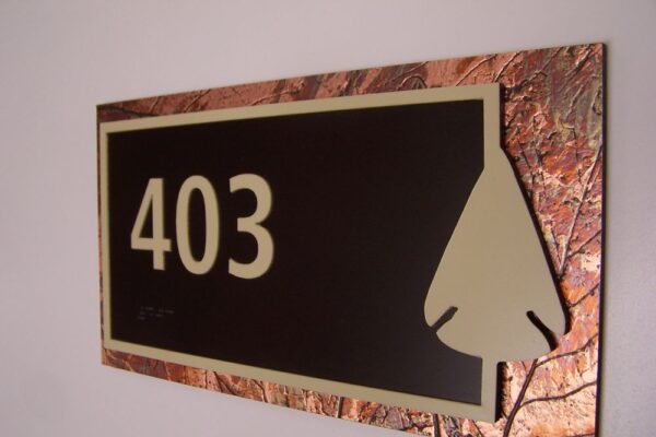 A rectangular sign on a wall displays the number "403" in large beige numerals on a dark brown background. The sign has a textured copper-colored border and includes a stylized leaf shape in the lower right corner.