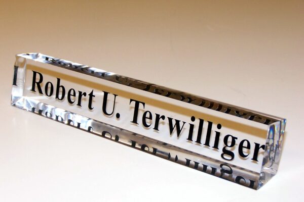 A rectangular glass nameplate with black text that reads "Robert U. Terwilliger" is displayed against a light background. The transparent glass reflects light, highlighting the crisp, clear text within.