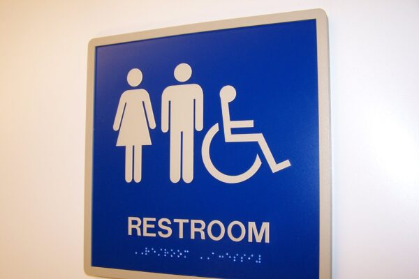 A blue restroom sign featuring white symbols for male, female, and wheelchair accessibility. The word "RESTROOM" is printed below the symbols, with additional text in Braille beneath it.
