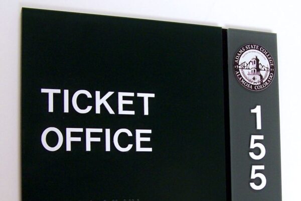 A b;lack and green sign mounted on a wall reads "TICKET OFFICE" in white uppercase letters, with "155" vertically aligned on the right side. The upper right corner features a circular emblem with text and a small building illustration. Braille is included below the words.
