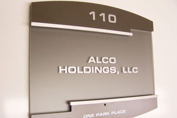 A door sign with the number "110" at the top. Below, it reads "ALCO HOLDINGS, LLC" in white capital letters. At the bottom of the sign, it says "ONE PARK PLACE." The sign has a modern style with a metallic finish and white text.
