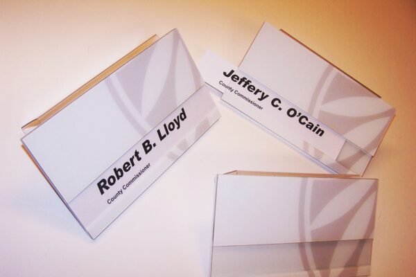 Three nameplates are displayed against a white surface. Each nameplate is clear with black text showing names: "Robert B. Lloyd," "Jeffery C. O'Cain," and another with the name blank. The nameplates are positioned upright in holders.