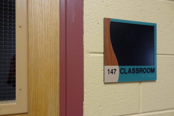 A door with a window panel on the left side and a classroom sign on the right. The sign is marked "147 CLASSROOM" with a decorative black and wood-colored design at the top.