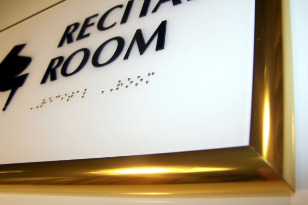 A close-up of a white sign with black text that reads "RECITAL ROOM" in all capital letters. Below the text, there is Braille. The sign is bordered by a gold frame.