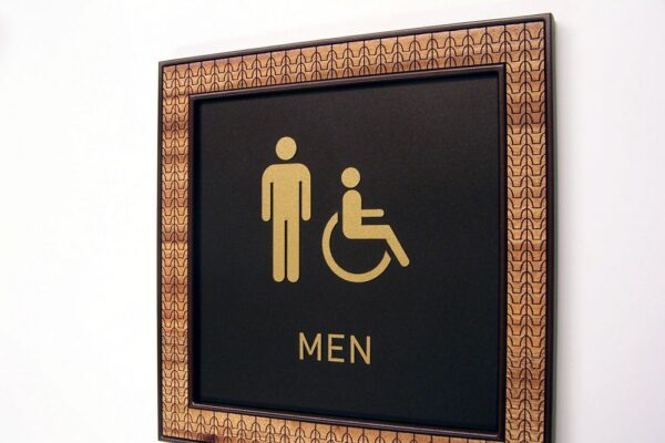 A bathroom sign indicating a men's restroom, featuring gold icons of a standing man and a person in a wheelchair on a black background. The sign is surrounded by a decorative, patterned frame.