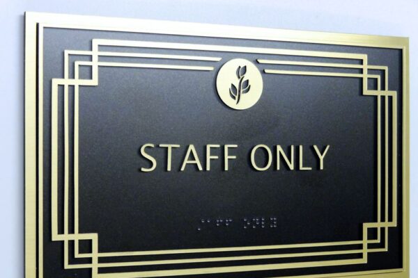 A black sign with gold trim and a gold emblem of leaves at the top center reads "STAFF ONLY" in gold uppercase letters. Below the text, there are tactile braille characters for accessibility. The sign is mounted on a light-colored wall.