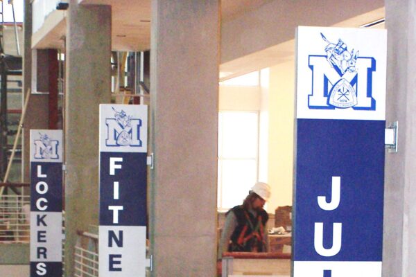 Banners hang vertically from columns inside a multi-level building, labeled "LOCKERS," "FITNESS LAB," and "JUICE LAB," respectively. Each banner features the same blue and white color scheme, with the letter "M" and a stylized knight's helmet logo at the top.