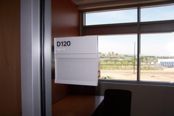 A sign with the text "D120" and a name is mounted on a wall, marking an office door. The office has a window with a partially closed blind and a view of an outdoor landscape. An empty desk and office chair are visible inside the office.