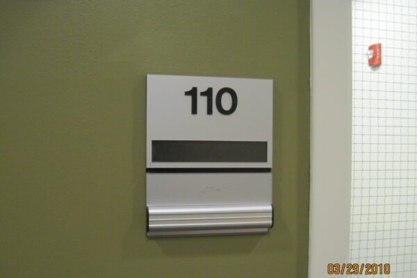 A sign on a wall displays the number "110" in bold black font. Below the number, there is a blank black stripe and a horizontal strip with braille. A partial white door frame with a mesh window is visible on the right.