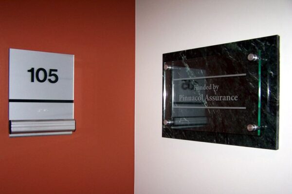 A hallway sign with the number "105" on the left. To its right, a black plaque mounted on the wall displays the text "Funded by Financial Assurance." The background features a two-toned wall, with one side painted orange and the other white.