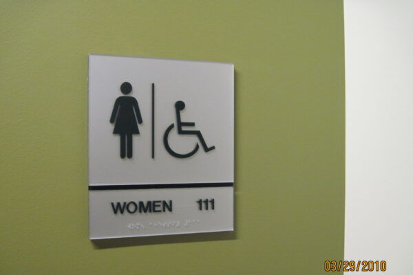 A sign on a green wall indicating a women's restroom with accessibility features. The sign includes icons of a woman and a wheelchair, the word "WOMEN," and the number "111" as well as braille text underneath.
