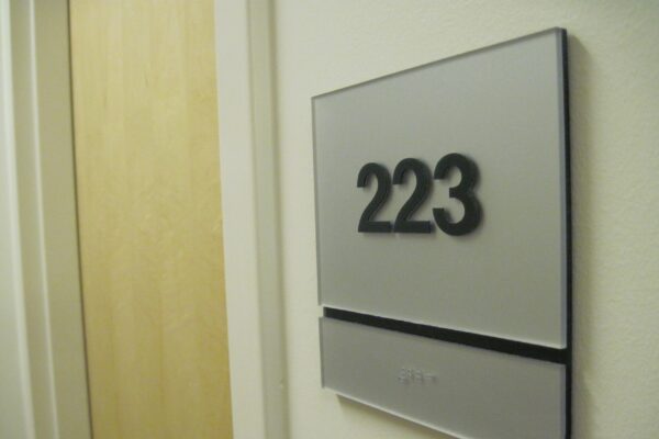 Sign displaying room number 223 mounted on a wall near a light wooden door in a building hallway. The sign has a metallic appearance with bold black numbers and a narrow horizontal strip underneath.
