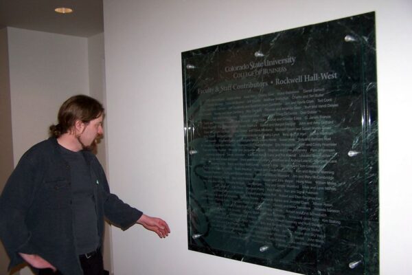 A person with long hair wearing a dark jacket and shirt points to a polished stone plaque mounted on a white wall, which appears to contain inscribed names or text.