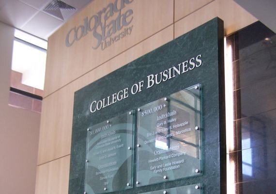 An interior display at a university's College of Business features a tall, green marble-like plaque with glass panels listing names. The backdrop includes wooden paneling and part of a stairwell. Natural light illuminates the area.
