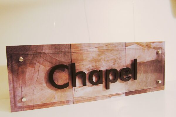 A sign mounted on a wall reads "Chapel" in bold, raised lettering. The background of the sign has a subtle, earthy-toned pattern with a rectangular shape and secured at the corners with visible screws. The overall look is modern and clean.