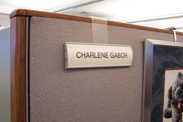 A nameplate reading "CHARLENE GABOR" is affixed to a cubicle wall. The surroundings include a grey fabric partition and part of a picture frame on the right side. The nameplate is held up by a clear hanger over the edge of the cubicle wall.
