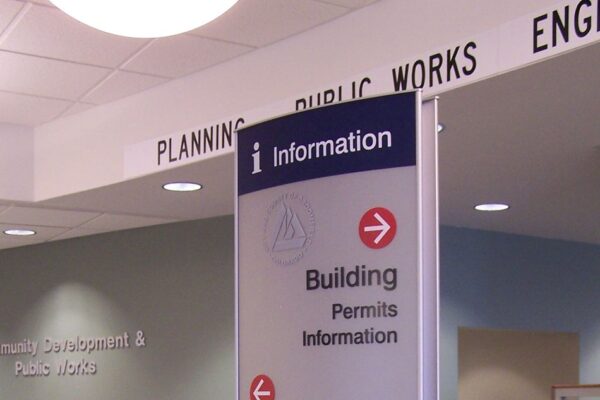 A directional sign in an office lobby displaying information for building permits, planning, engineering, CIP, GIS, and public works. The sign has arrows indicating directions. Surrounding the sign are desks, papers, and partial views of office equipment.