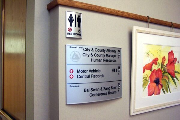 A hallway wall displays directional signs. A restroom sign with male and female icons is at the top. Below it, a sign lists several departments: the Attorney, Manager, Human Resources, Motor Vehicle, Central Records, and a conference room. A framed flower artwork hangs nearby.
