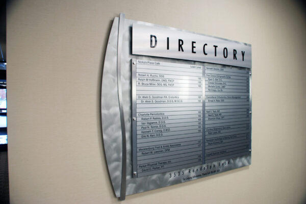 A metallic building directory sign is mounted on a light-colored wall. The sign is sleek and modern, with the word "DIRECTORY" prominently displayed at the top. Below are rows of engraved names and suite numbers, organized in a clear, easy-to-read format.