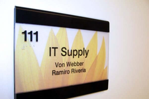 A door sign with a sunflower design shows the text "111 IT Supply" and lists two names: "Von Webber" and "Ramiro Riveria." The sign is mounted on a white wall.