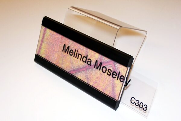 A nameplate with the text "Melinda Moseley" is displayed on a clear stand. The nameplate has a multicolored background with a mix of pink, yellow, and purple hues, and is bordered by black strips at the top and bottom. The label "C303" is visible beneath the stand.