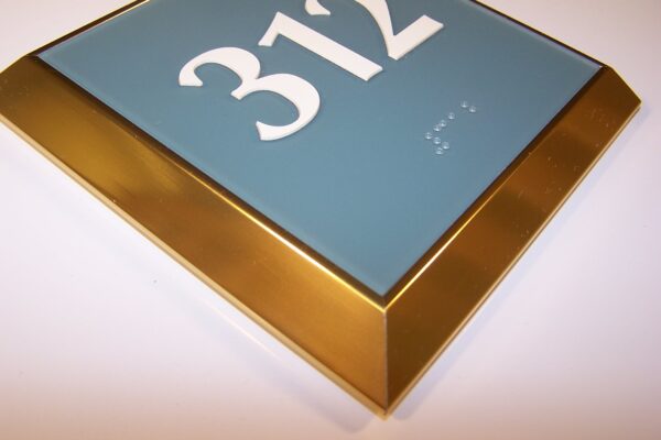 A close-up of a rectangular room sign displaying the number 312 in large white characters against a blue background. The sign has a gold border with a beveled edge and includes Braille dots below the numbers. The sign is mounted against a white surface.