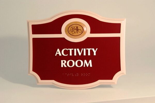 A red and white sign with an ornate shape, displaying the words "ACTIVITY ROOM" in bold white letters. The top part of the sign features a small circular emblem with an illustration of a flower inside it.