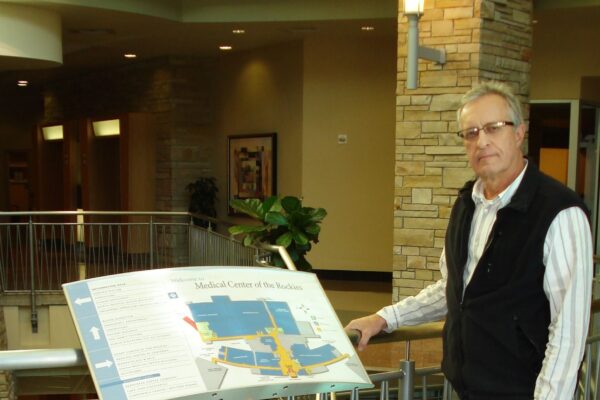 A person standing indoors next to a large directory sign mounted on a railing. The directory displays a map and text. The person is dressed in a striped shirt, dark vest, gray pants, and black shoes, and is looking towards the camera. The background includes a staircase.