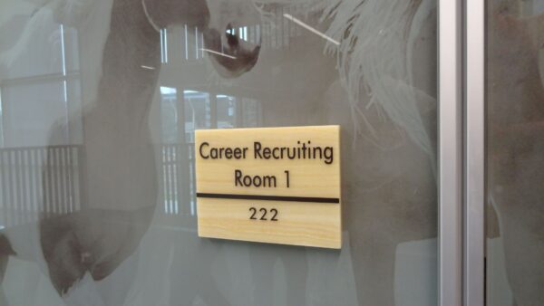 A beige sign on a glass door reads "Career Recruiting Room 1" with the number "222" beneath it. The background shows a corridor with glass walls and reflective surfaces.