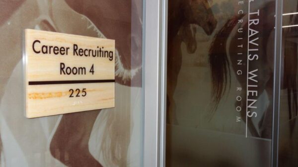A close-up photo of a wooden door plaque that reads "Career Recruiting Room 4," with the number "225" below it. The door has a frosted glass panel with partial text visible, including a part of the name "TRAVIS WIENS.