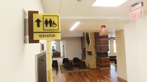 A hallway with a yellow elevator sign indicating accessible facilities, accompanied by a directional arrow. The corridor features wooden flooring, comfortable seating, a fireplace, and a "EXIT" sign. Natural light enters through a window at the end of the hall.